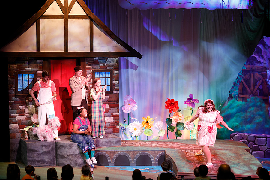 Photo of several people on a stage decorated with a house and a garden. One person is wearing white overalls and operating a pink dog puppet.