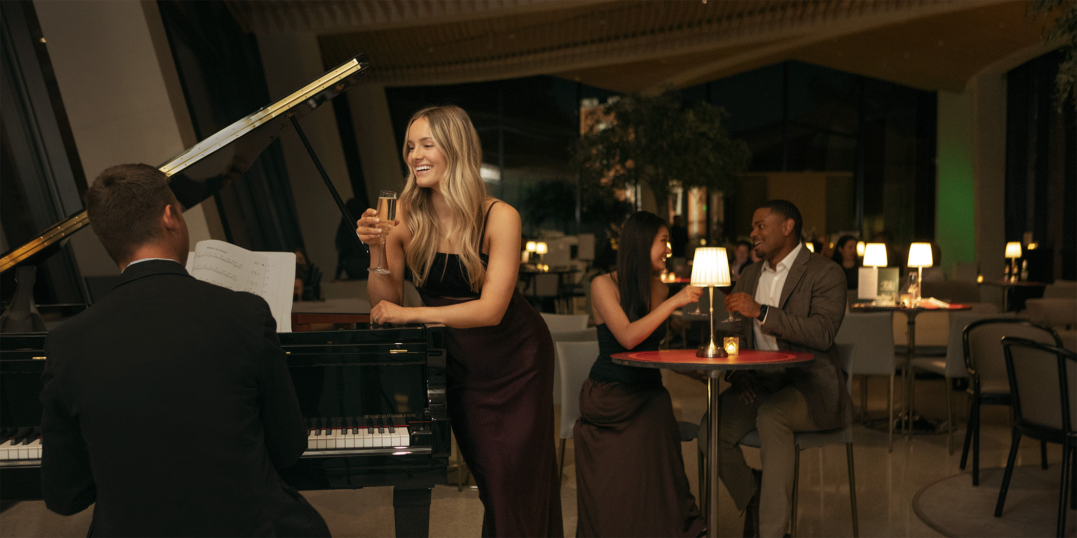 Photo of a woman holding a glass of champagne and leaning on a grand piano played by a seated man. People are seated at small tables with lamps in the background.