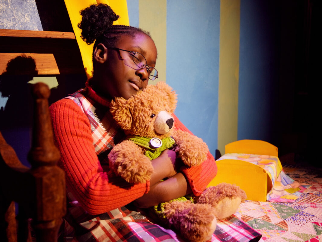 Photo of a girl performing on stage seated on a bed and holding a teddy bear.