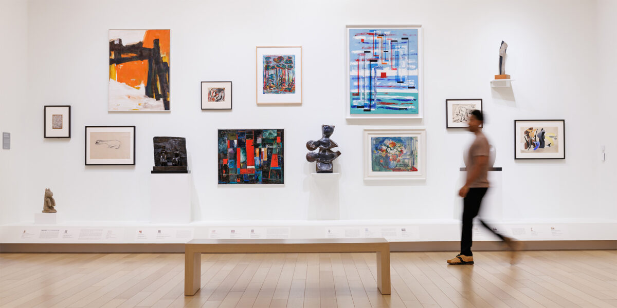 Photo of a man walking in front of a bench next to a wall of framed art in an art gallery.
