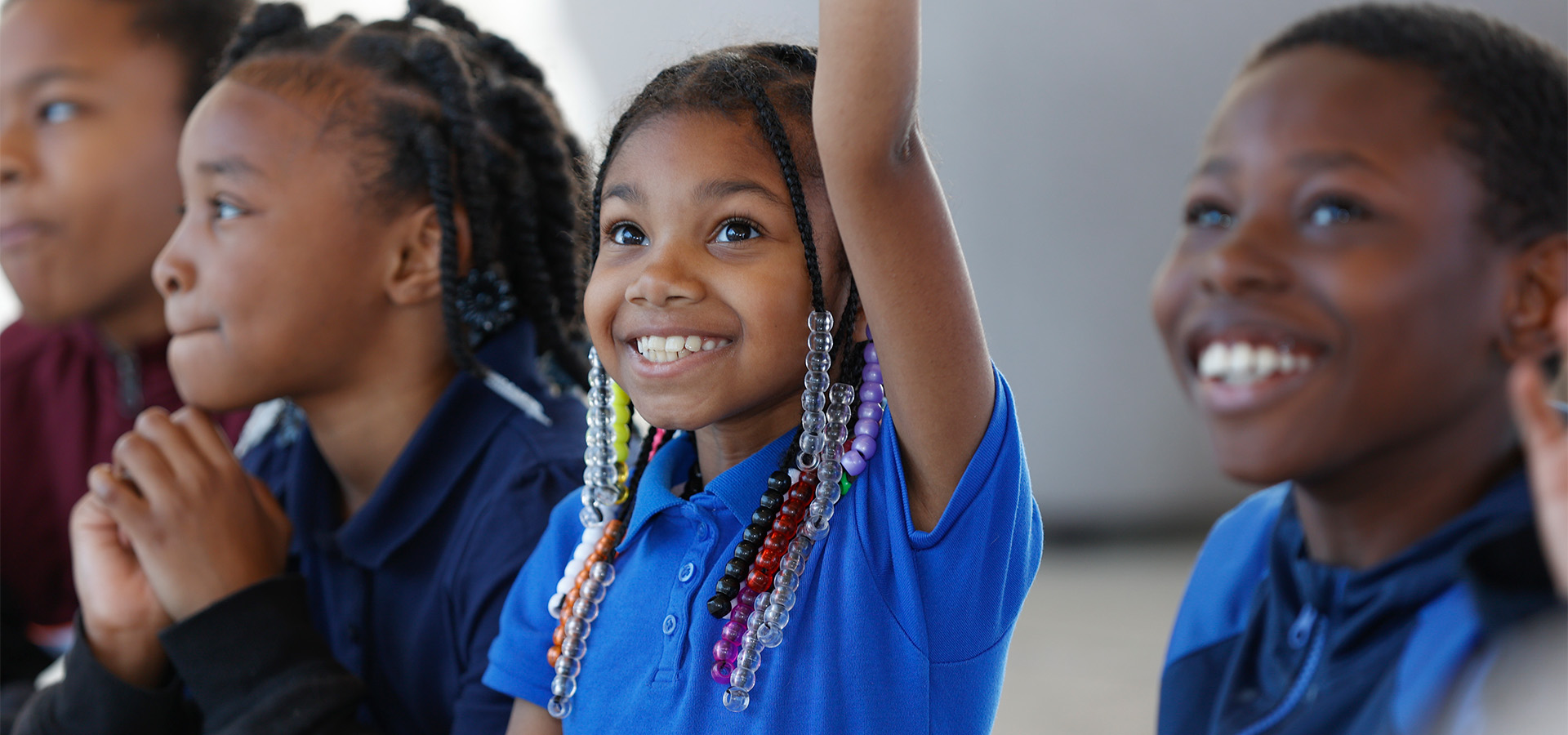 Close up photo of several young children smiling. One girl has beads in her hair and her hand raised.