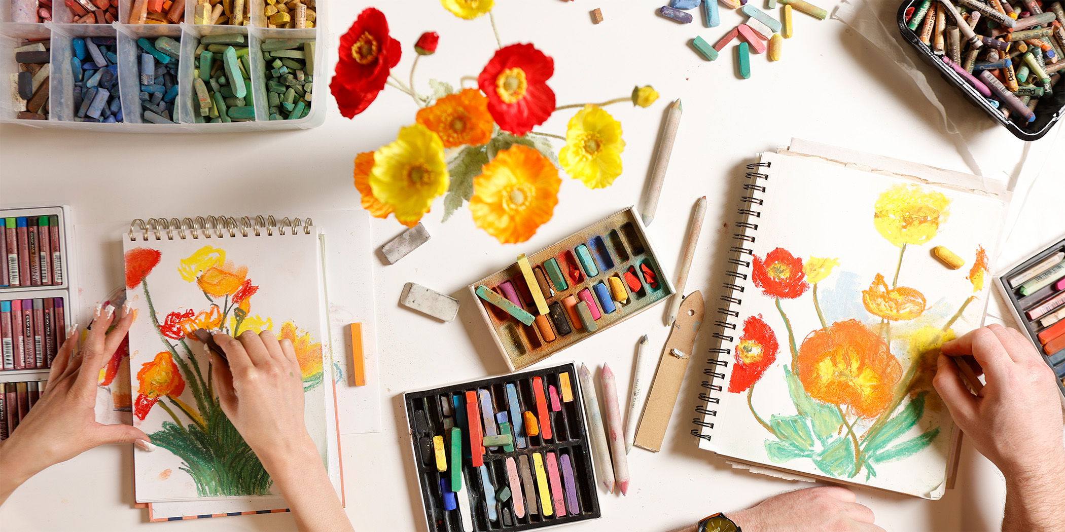 Overhead photo of two hands drawing flowers on drawing pads next to a floral arrangments and boxes of color pastels.
