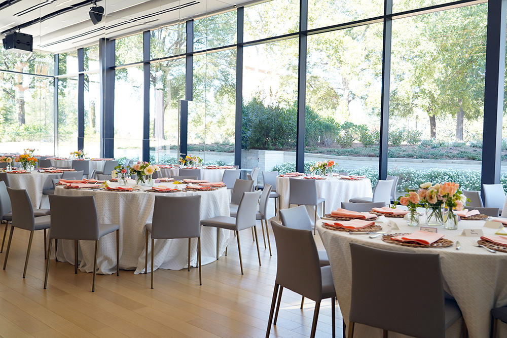 Photo of a room with wood panel flooring and floor to ceiling windows decorated with round tables covered with white tableclothes and orange napkin place settings next to gray chairs.