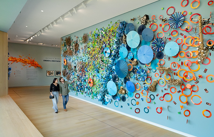 Photo of a man and a woman walking in an art gallery with a colorful art installation on a blue wall next to them.