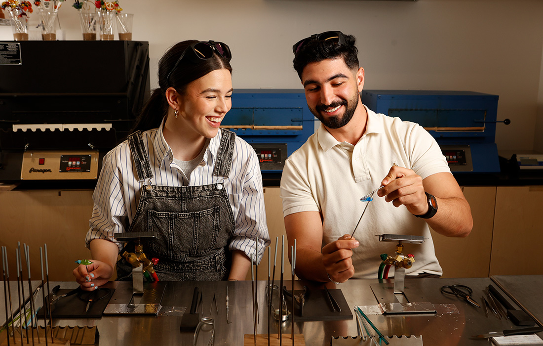Photo of a woman and a man seated and smiling while looking at a small handmade glass object.