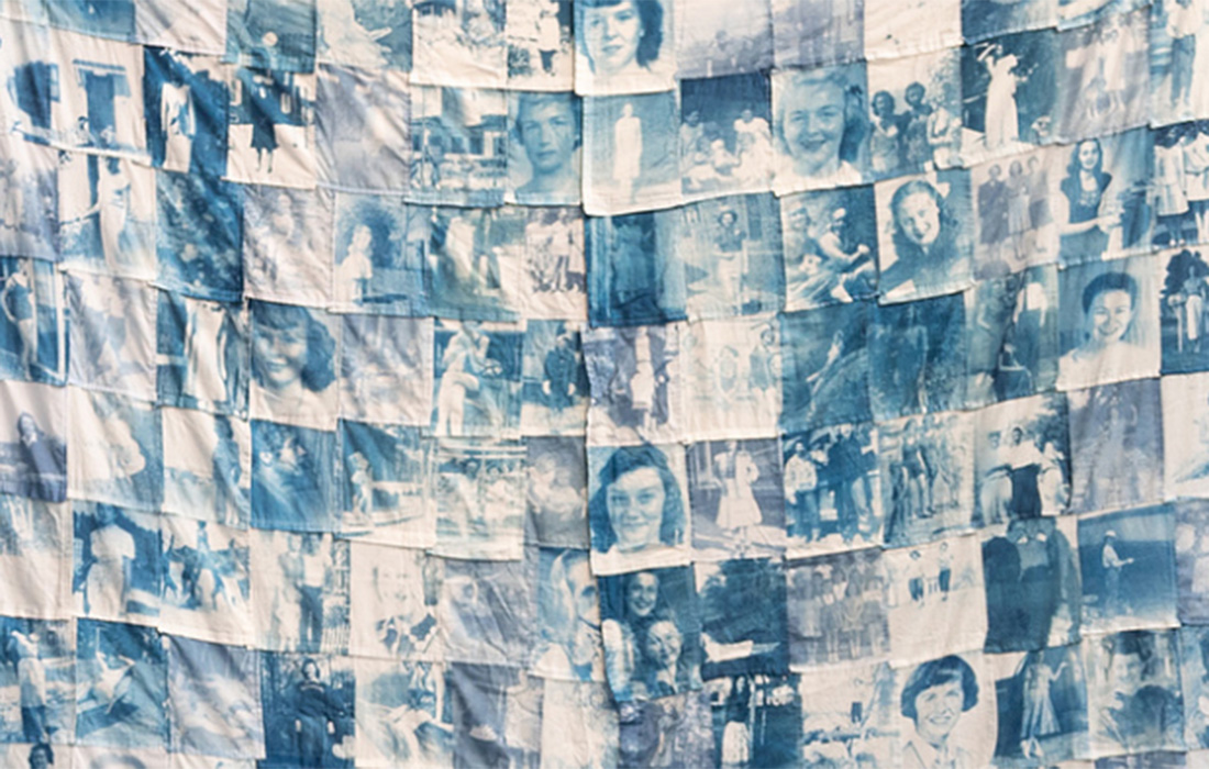 Photo of a tapestry quilt made of small fabric sections depicting people's faces in blue ink.