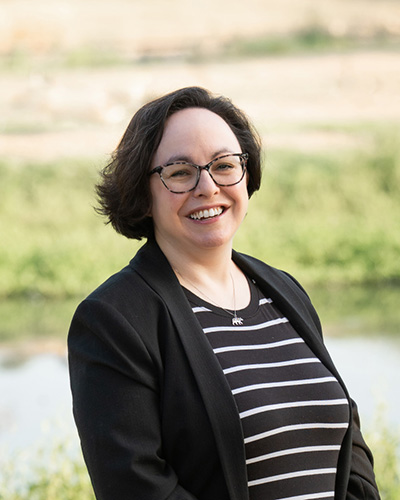 Headshot of a a woman with short brown hair wearing glasses, a black blazer, and a black and white striped blouse photographed outdoors.