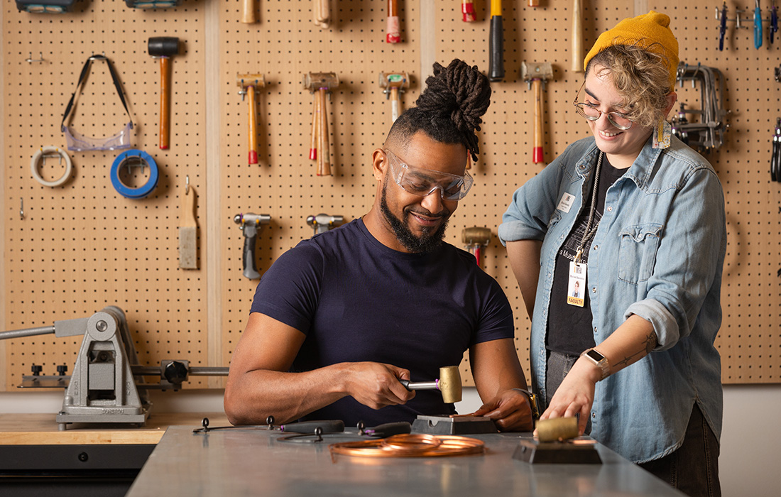 Photo of a seated man hammering a piece of metal while an instructor wearing a badge and a yellow beanie stands next to him looking on.