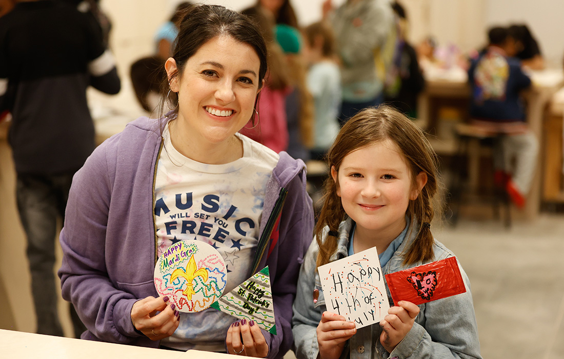 Photo of a mother and daughter seated and smiling in an art studio while holding up small drawings.