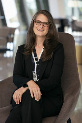 Photo of a woman with long brown hair wearing glasses and a black blazer over a black and white patterned blouse while seated in a dark gray chair.