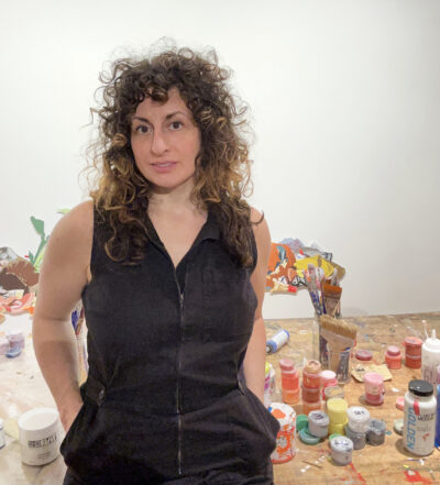 Photo of a woman with medium-length curly brown hair wearing a sleeveless black leather jumpsuit. Behind her are bottles of paint on a hardwood floor.