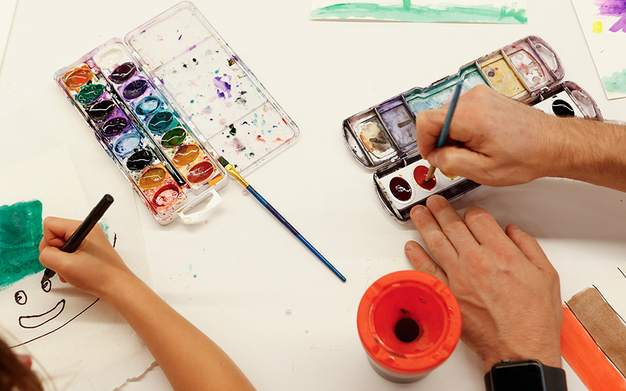 Overhead photo of adult hands painting with watercolors next to child hands drawing a smiley face using a black marker on a white surface.