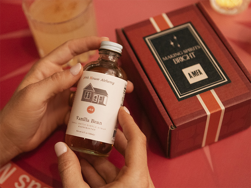 Photo of two hands holding a bottle of vanilla bean syrup next to a red box that reads Making Spirits Bright above an AMFA logo.