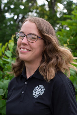 Headshot of a woman with short brown wearing wearing glasses and a black collared shirt with a white logo.