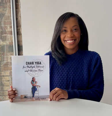 Photo of a woman wearing a dark blue sweater seated at a white table and smiling at the camera while holding a book.