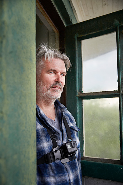 Photo of a man with short gray hair and stubble wearing a plaid shirt and standing in a doorway.