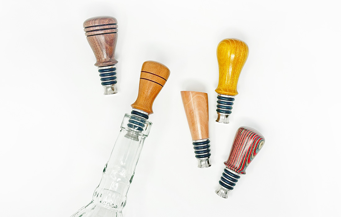 Photo of an empty glass bottle with a handmade, wood-turned bottle stopper inserted in the top. Four other stoppers in varying designs are laying on a white surface next to the bottle.