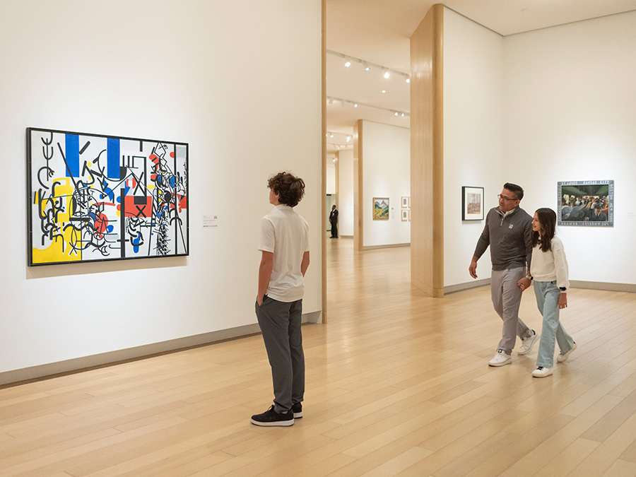 Photo of a boy looking at an abstract painting on a wall in an art gallery. A man in the background is holding hands and walking with a young girl.