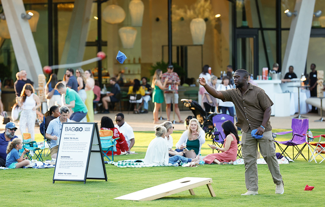 Photo of people on the Event Lawn at AMFA. In the foreground is a man playing baggo surrounded by people sitting on blankets and talking. In the background are people standing and holding drinks.