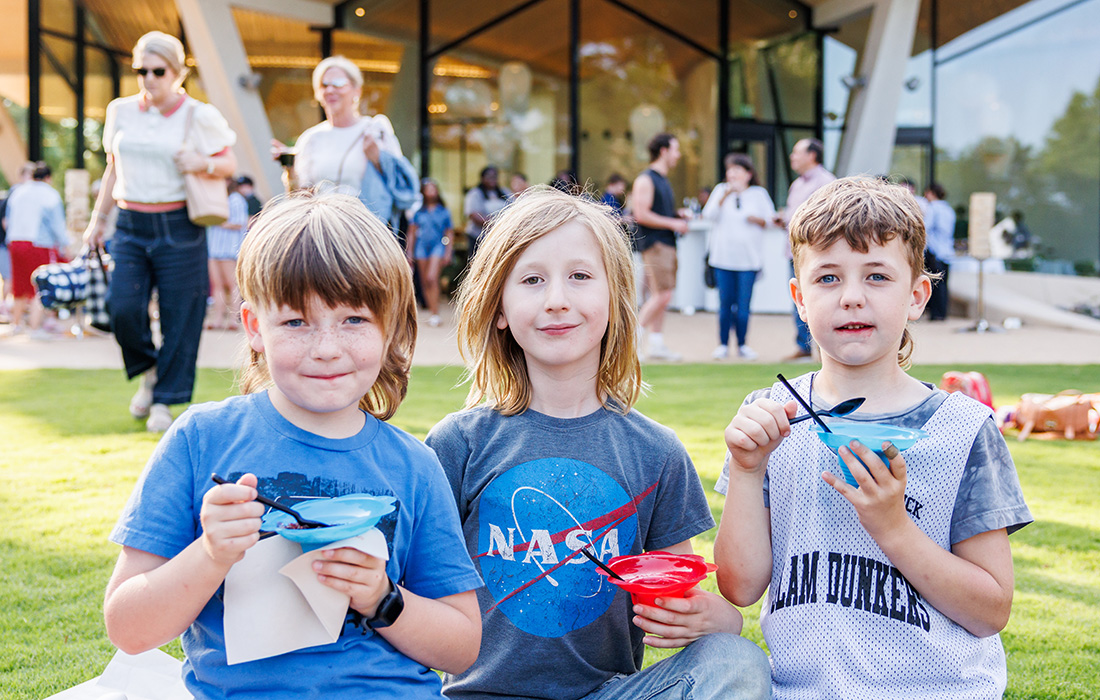 Photo of three young boys seated outside on the Event Lawn at AMFA smiling and holding bowls containing a frozen dessert.