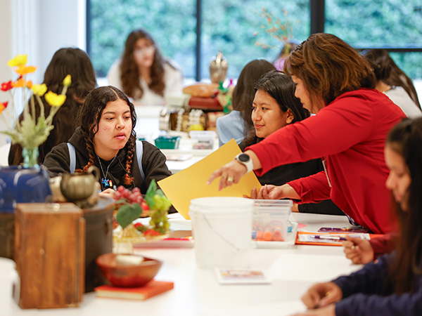Photo of two teenage girls sitting at a table in an art studio while a woman standing next to them gesturing toward a piece of paper.