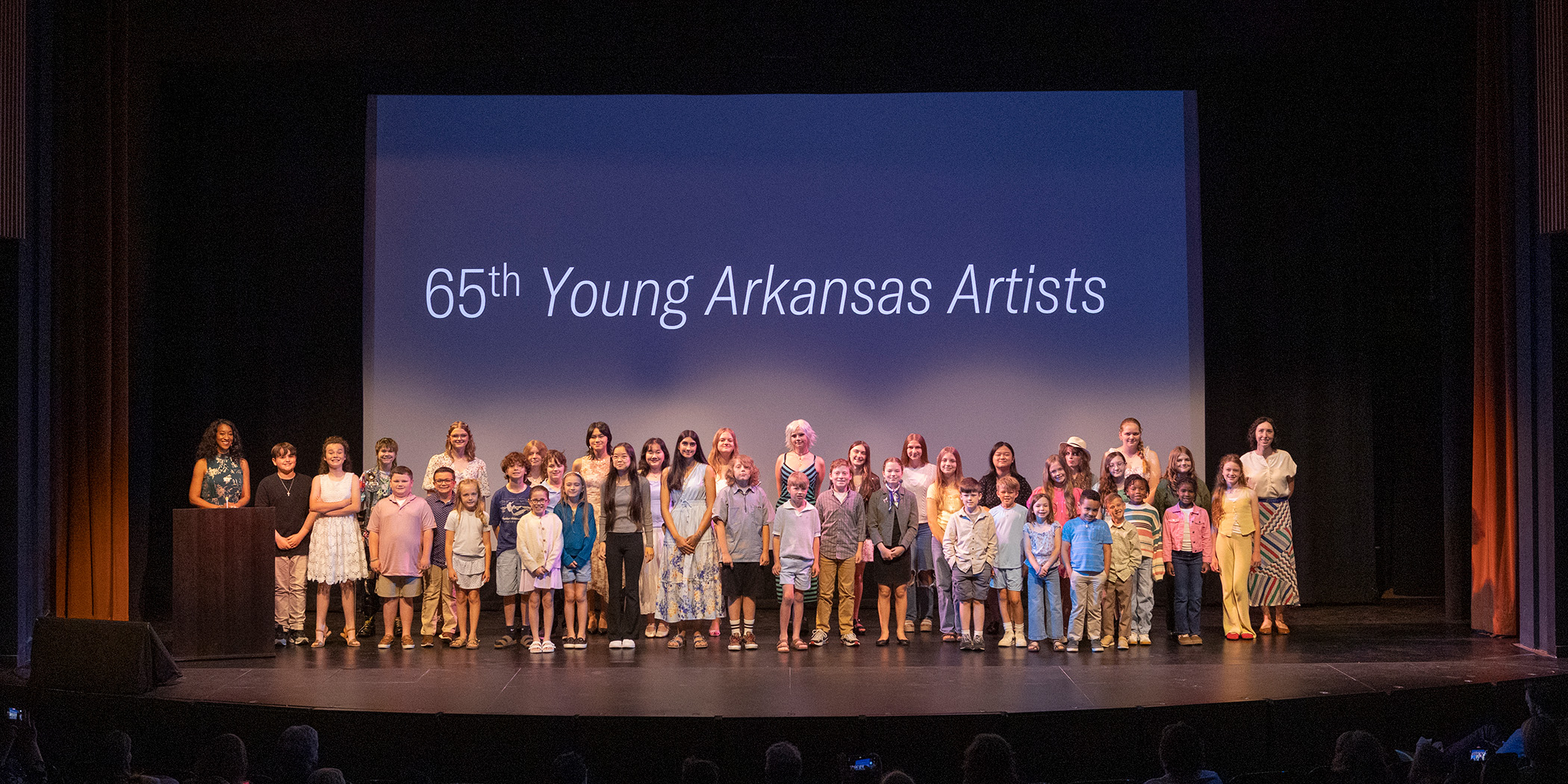 Photo of a group of young people posed together and smiling on a stage in front of a screen that says 65th Young Arkansas Artists.