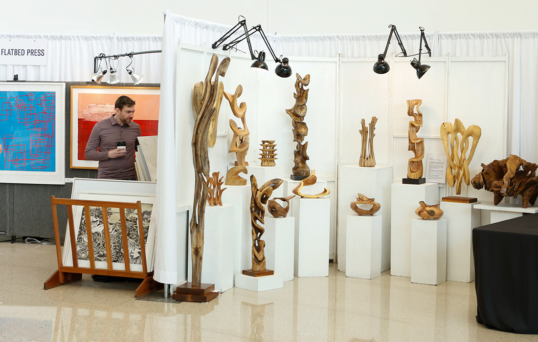 Photo of a main looking at paintings in an artist booth next to a booth containing wooden sculptures at an art market.