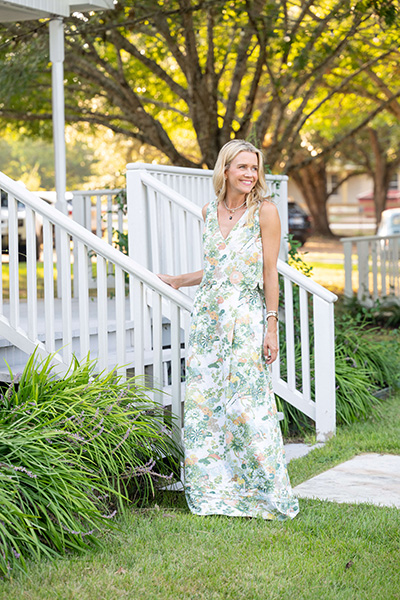 Photo of a woman smiling and wearing a floral dress posed on a green lawn in front of a porch with a white railing.