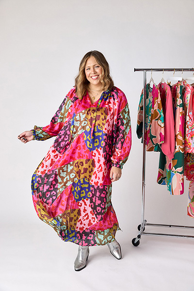Photo of a woman smiling and wearing a floral dress posed in front of a rack of clothing.