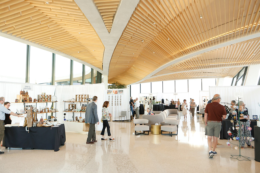 Photo of people walking amongst several booths at the AMFA Art Market in a room with a high curving ceiling and floor-to-ceiling windows.