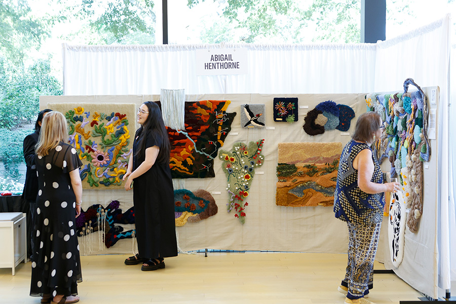 Photo of several women looking at large fiber artworks hanging in a booth at the AMFA Art Market.