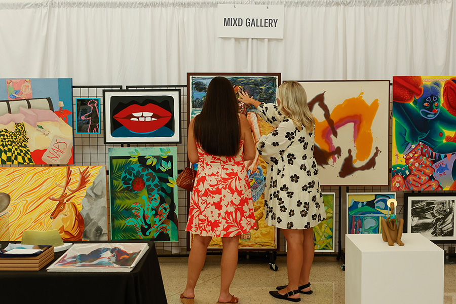 Photo of two women looking at paintings hanging in a booth at the AMFA Art Market.