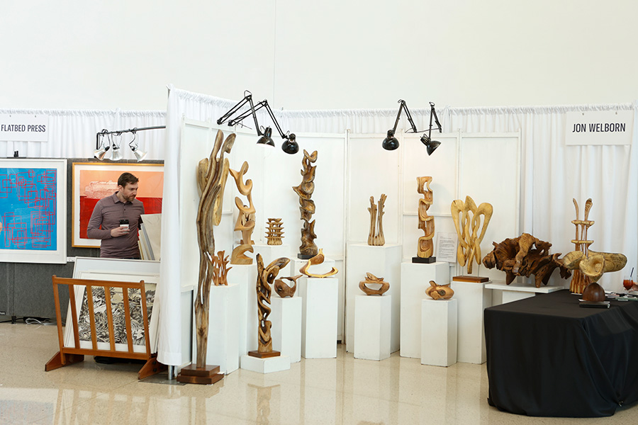 Photo of a man looking at paintings in a booth next to another booth filled with wooden sculptures at the AMFA Art Market.