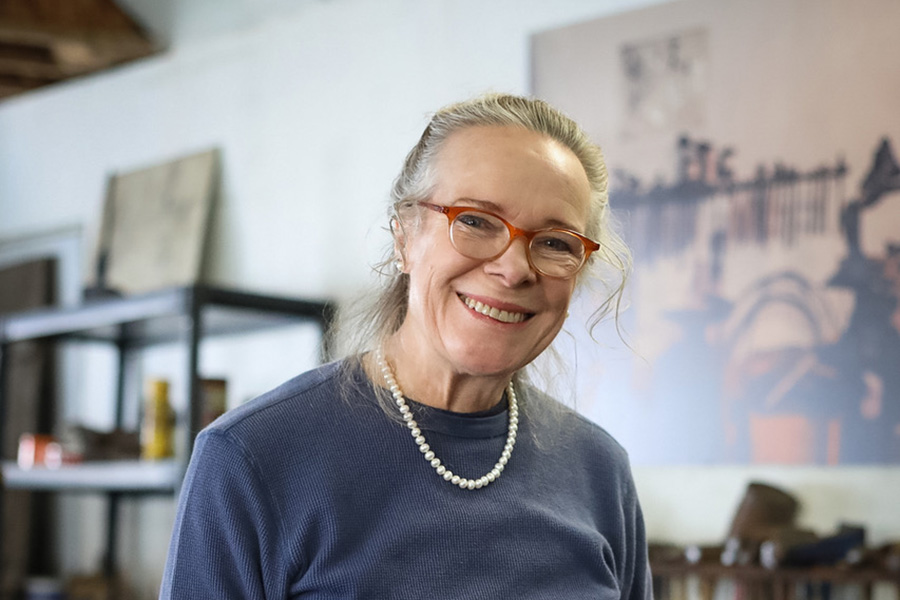 Photo of a woman with long gray hair pulled back into a ponytail. She is wearing a blue shirt, pearls, and red glasses.