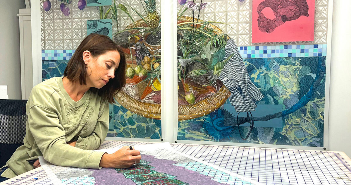 Photo of a woman with short brown hair wearing a light green shirt. She is seated and drawing on an artwork laid on a table in front of another large artwork on the wall behind her.