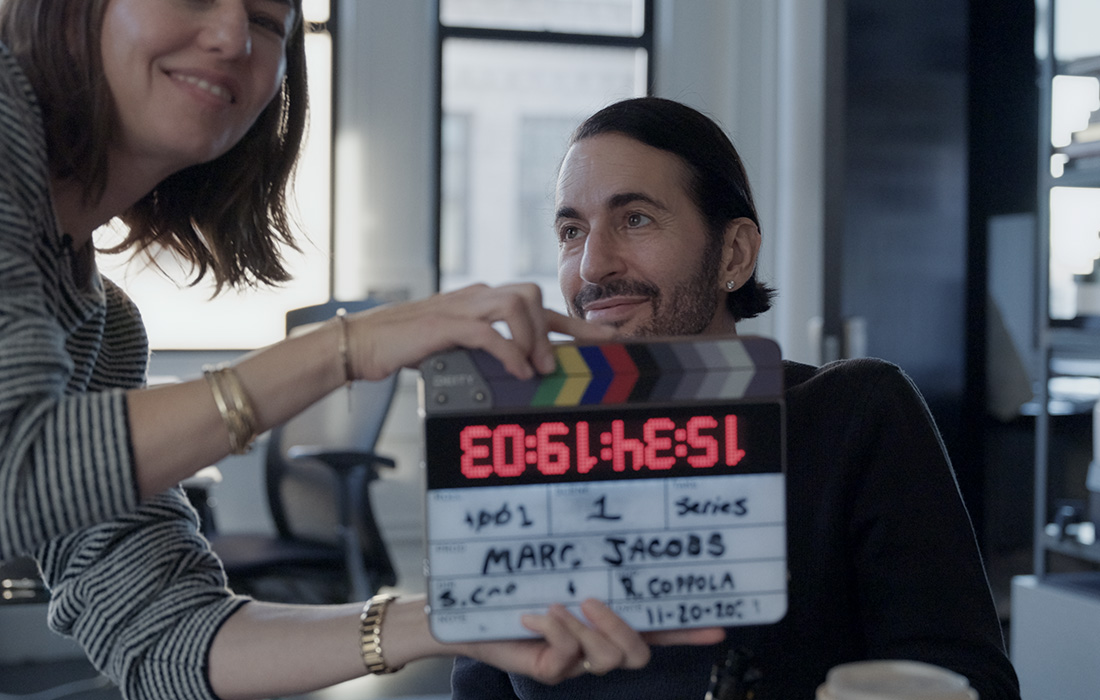 Film still depicting a woman with short dark hair smiling and holding a clapperboard in front of a seated man with dark hair and a mustache.
