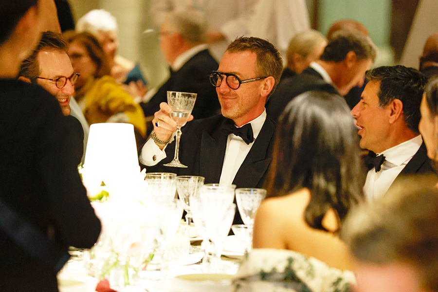 Photo of a group of people in formal wear seated around a table at a gala. A main in the center of the photo is wearing a tuxedo and black glasses and holding up a glass water goblet.