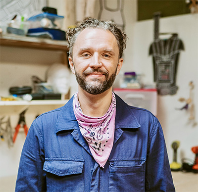 Headshot of a man with light brown graying hair and a beard wearing a blue shirt and pink bandana.