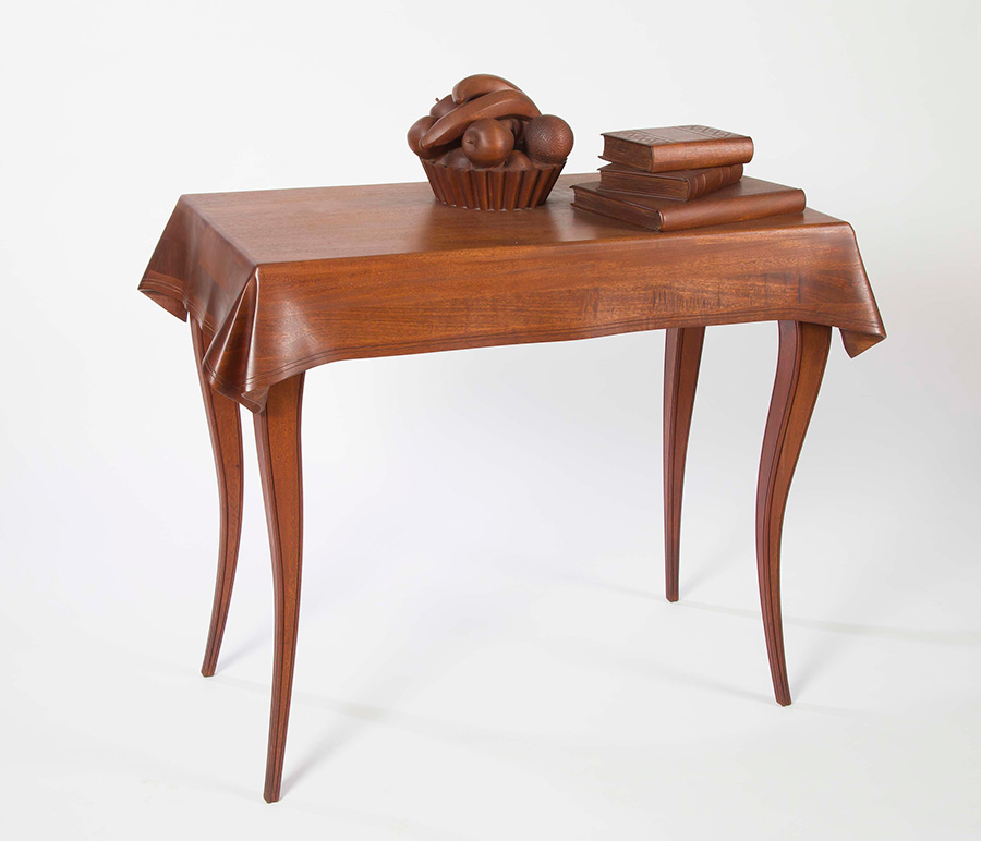 Photo of a wood sculpture in the shape of a table adorned with a bowl of fruit and a stack of books.