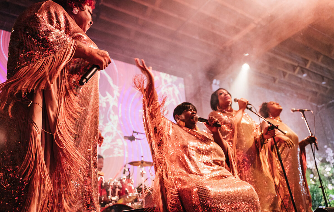 Photo of four women wearing pink sequin muumuus holding microphones and singing on a stage.