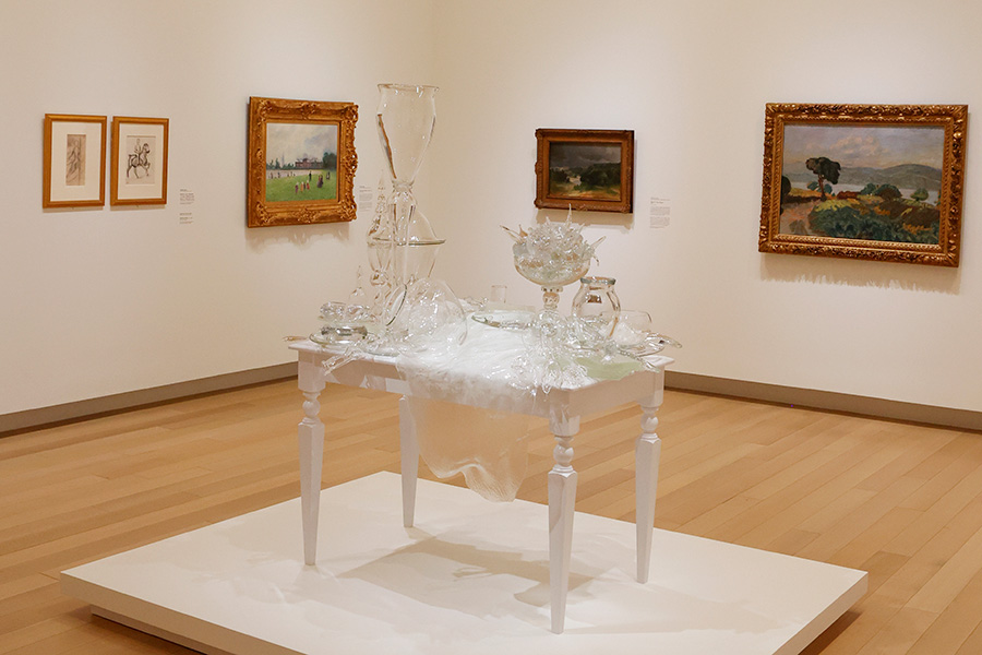 Photo of a glass sculpture in the shape of a table covered in food and other objects on a white platform in an art gallery.