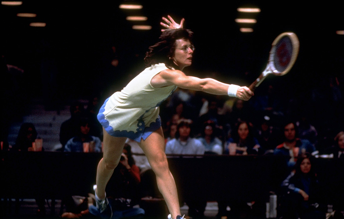 Photo of a woman with short brown hair wearing glasses and a white tennis dress mid-swing with a tennis racket in her hand in front of a crowd during a tennis match.