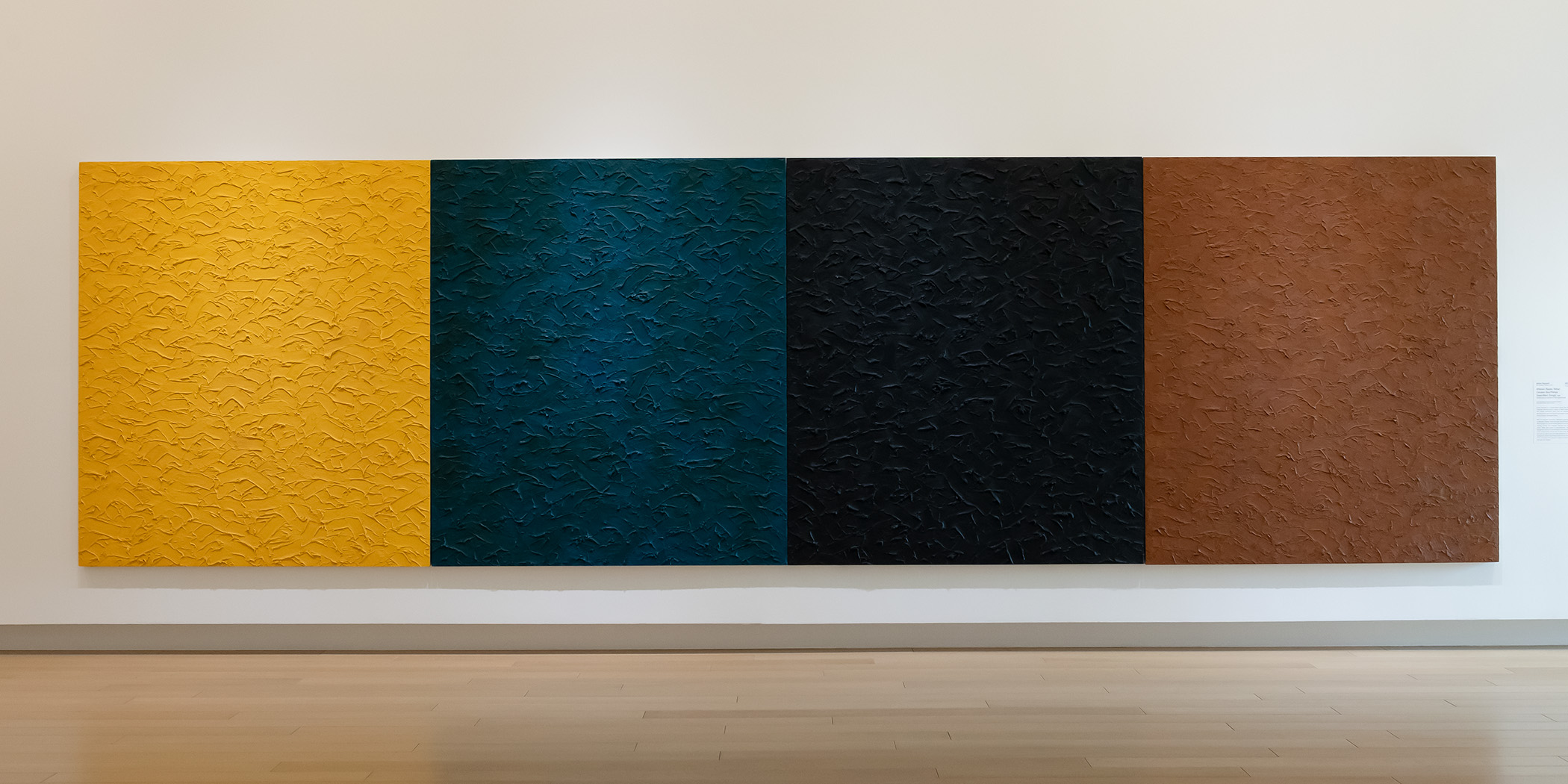 Photo of a large, four-paneled painting on a wall in an art gallery. Each panel has a distressed texture and is painted in a solid color.