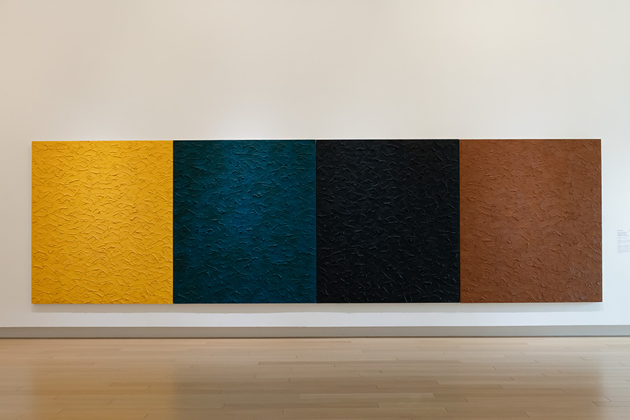 Photo of a large, four-paneled painting on a wall in an art gallery. Each panel has a distressed texture and is painted in a solid color.
