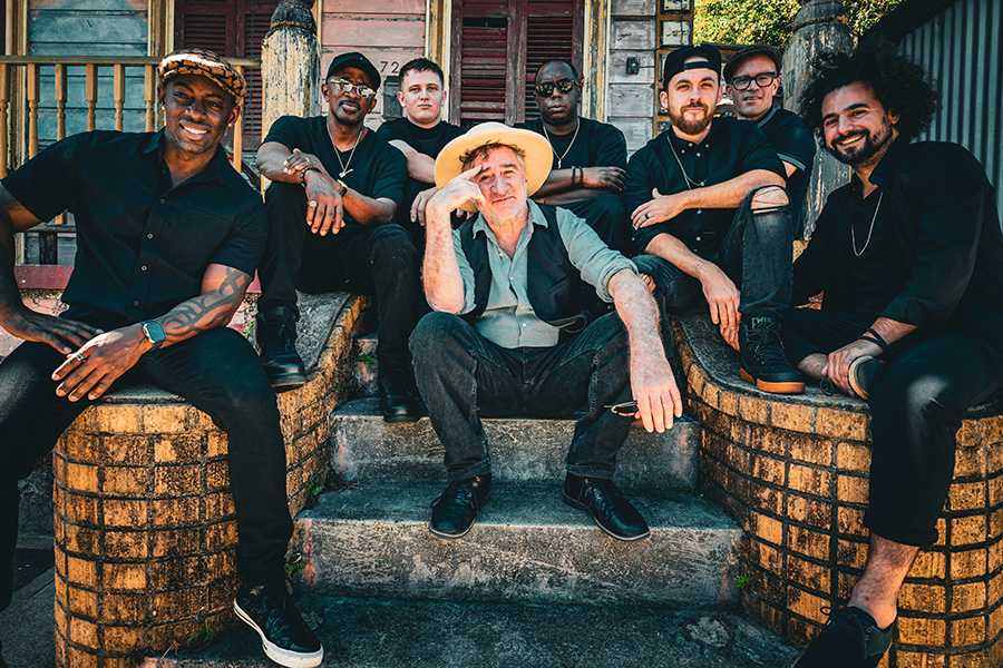 Photo of a white man wearing a vest, jeans, and a wide-brimmed hat sitting on concrete steps with a group of men wearing all black sit around him in various poses.