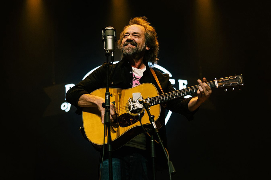 Photo of a man with long brown hair and a beard wearing a black jacket and holding an acoustic guitar while singing into a microphone on a dark stage.