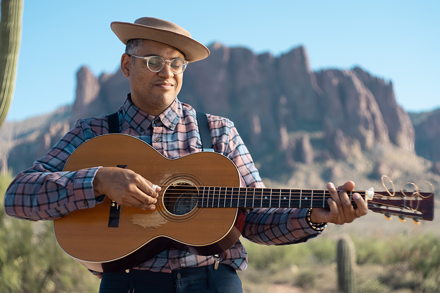 Photo of a man wearing a plain shirt, suspenders, and a brown hat holding an acoustic guitar in a desert next to a cactus.