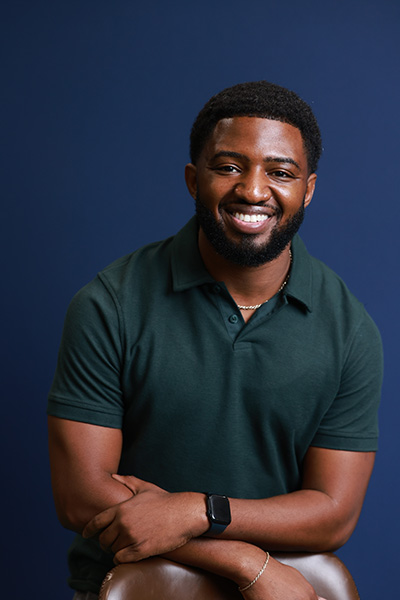 Photo of a man with short hair wearing a green polo and a black watch sitting with his arms crossed and smiling.