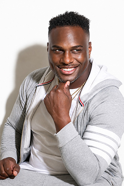 Photo of a man wearing a light gray tracksuit and white shirt posed smiling with his hand under his chin.
