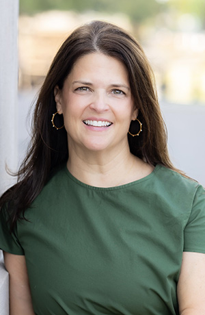 Headshot of a smiling woman with long brown hair wearing a green short-sleeved blouse and gold hoop earrings.